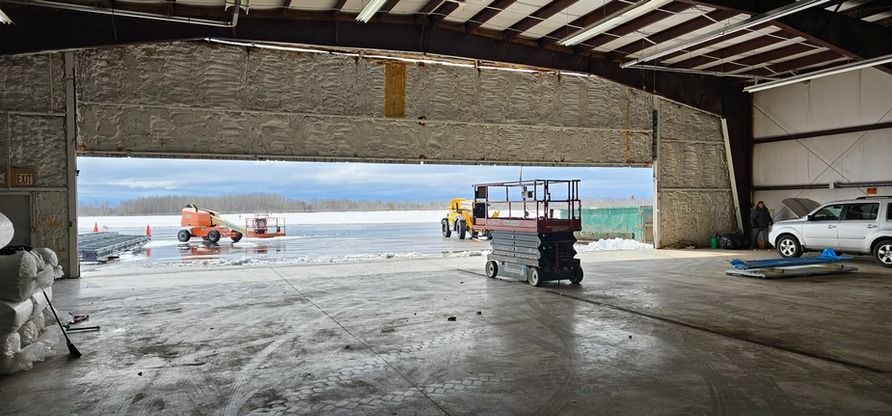 Inside a hangar, a lift and vehicles face a snowy landscape through an open doorway.