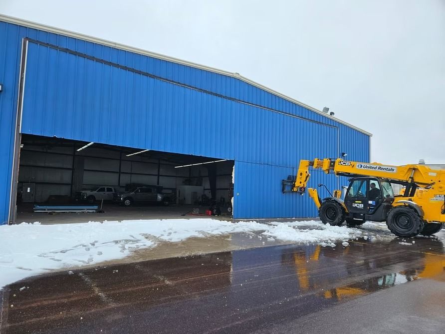 Blue hangar with partially open door, a construction vehicle outside on a snowy day.
