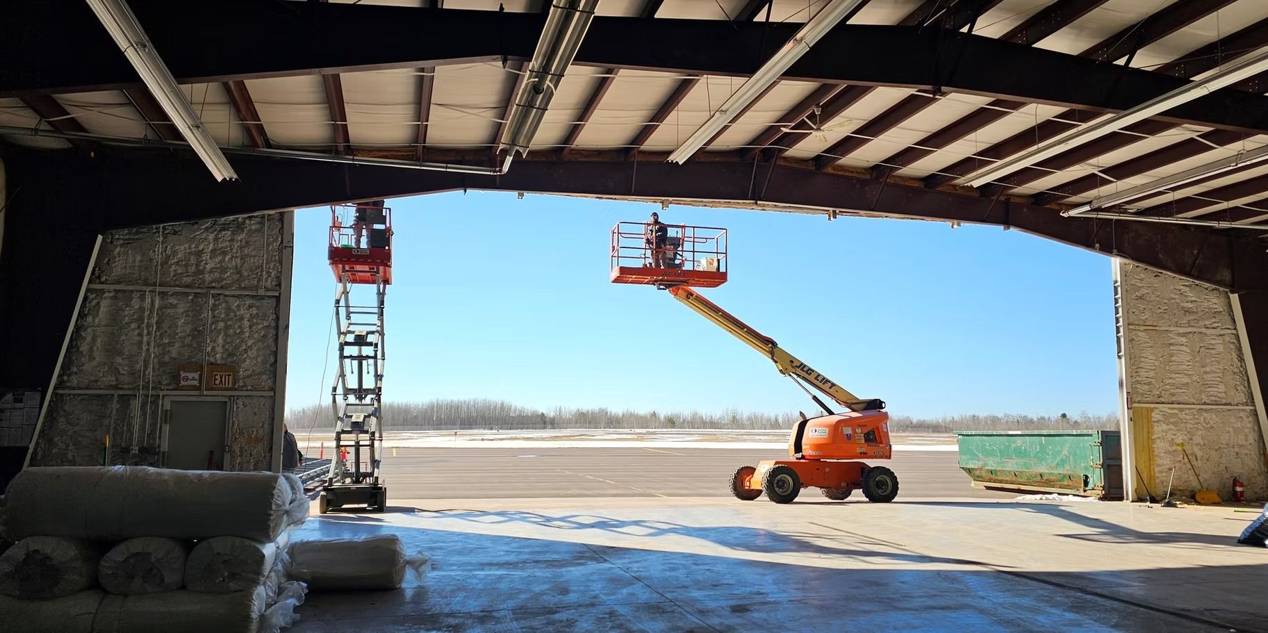 Workers on lifts inside a large building, possibly an airplane hangar, against a blue sky.