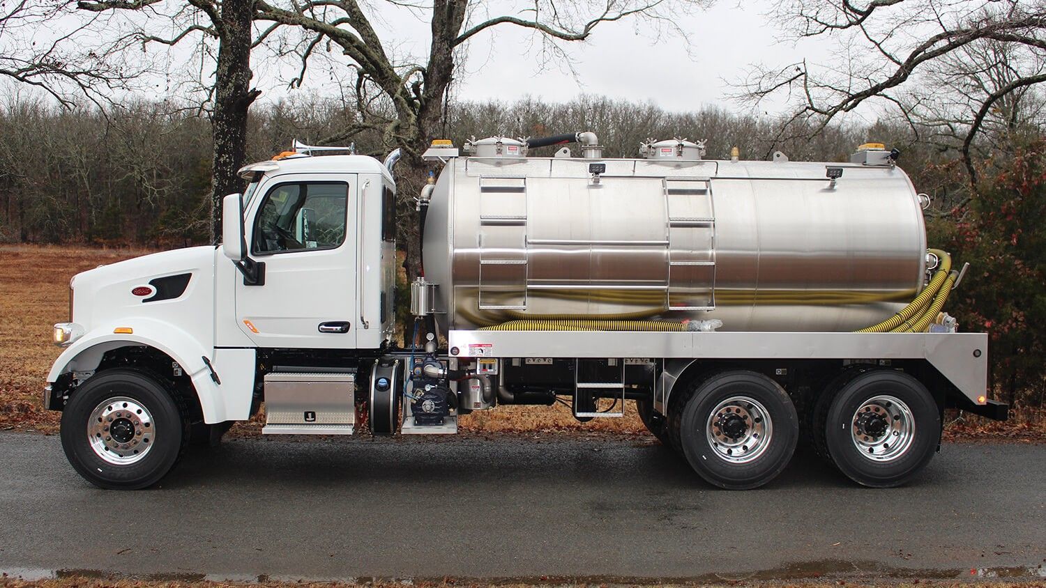 White septic tank truck on a road, parked in front of trees.