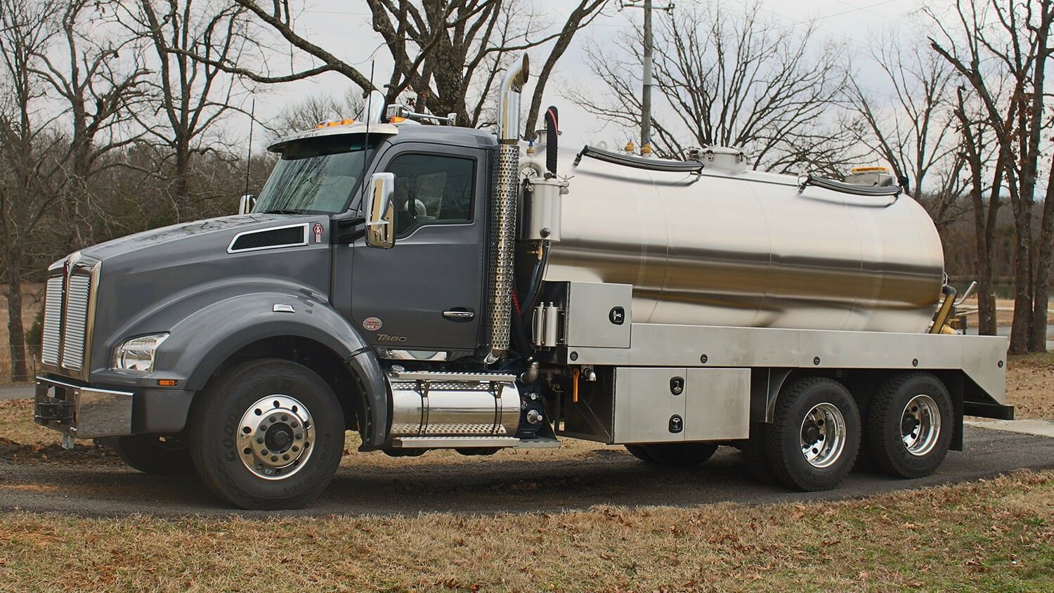 Grey tanker truck on a gravel road, stainless steel tank, trees in the background.