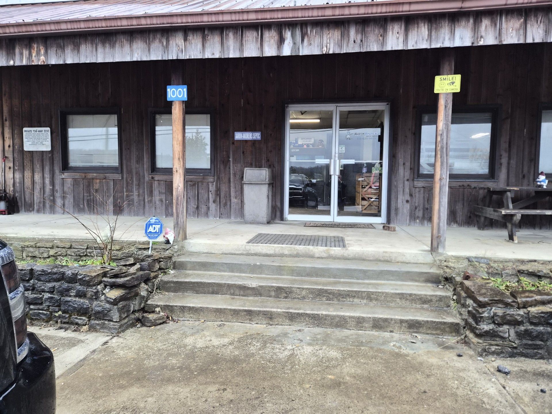 Exterior of a wooden building with a concrete entrance, glass doors, and a sign. Steps lead to the entrance.