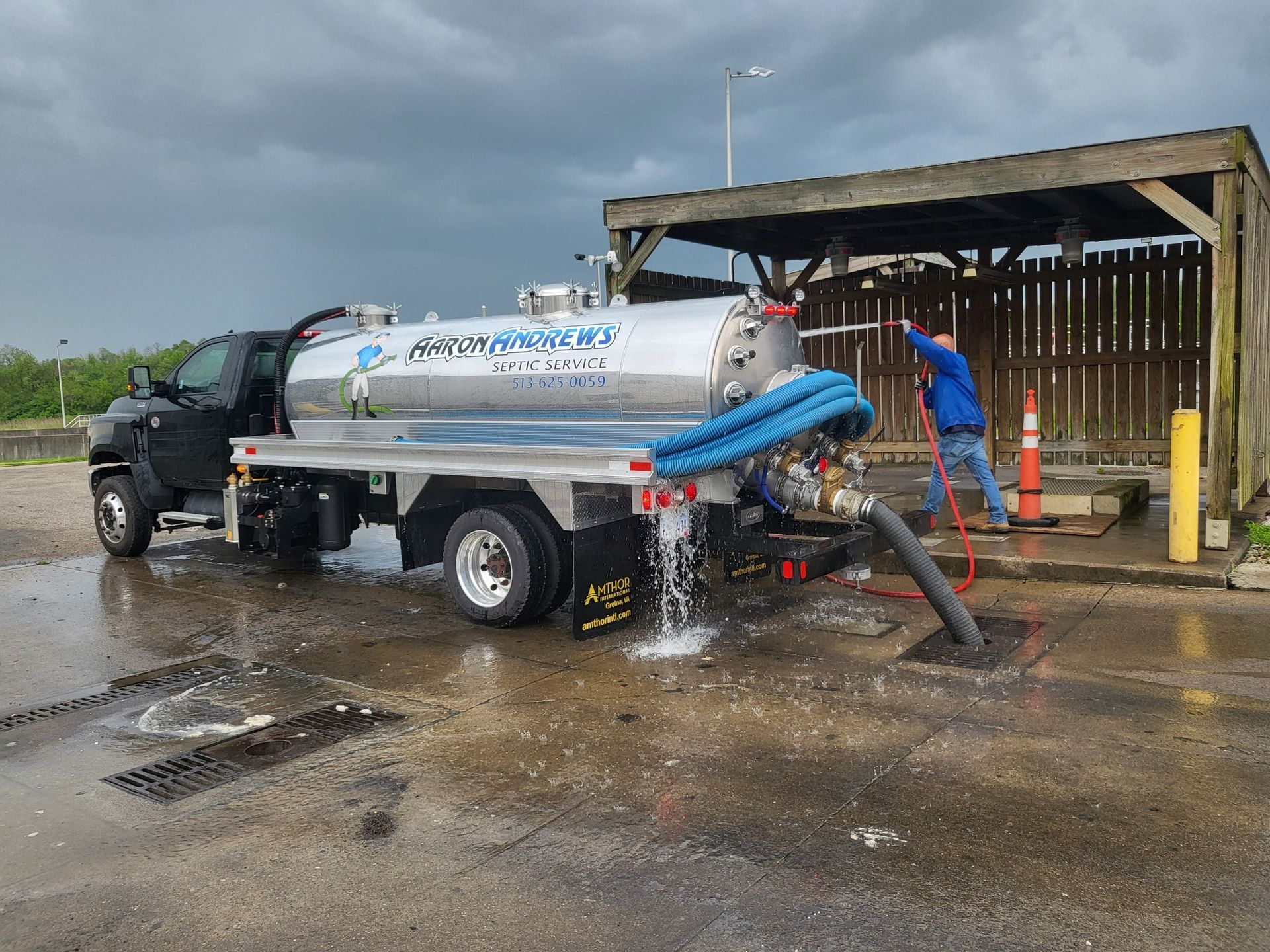 A black septic truck is discharging liquid waste. A man is holding a hose. Pavement is wet.