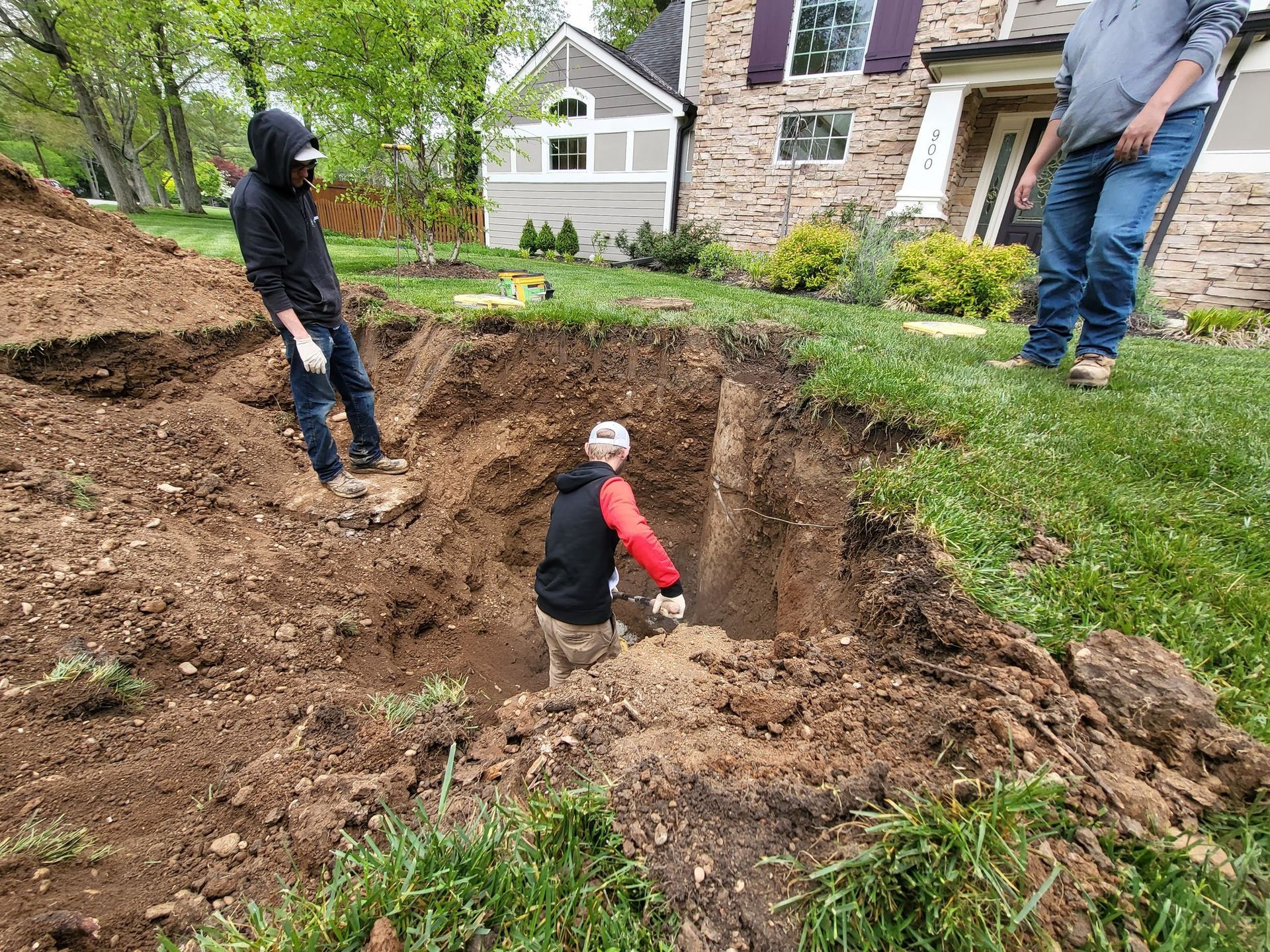 Three people dig in a large hole in a residential yard. Brown dirt surrounds the hole, and a house is visible in the background.