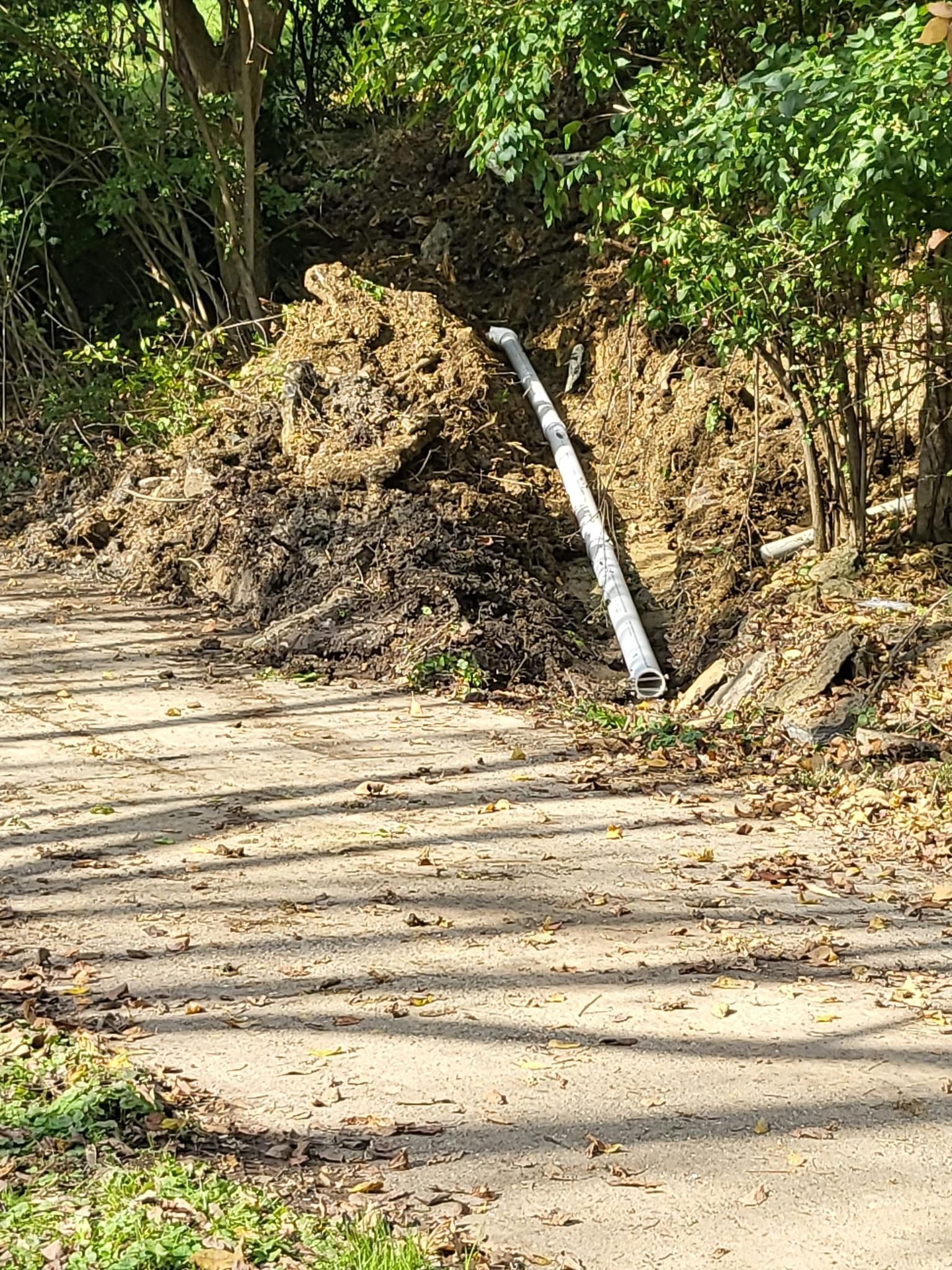 Pile of mulch with a pipe, next to a path in a wooded area.