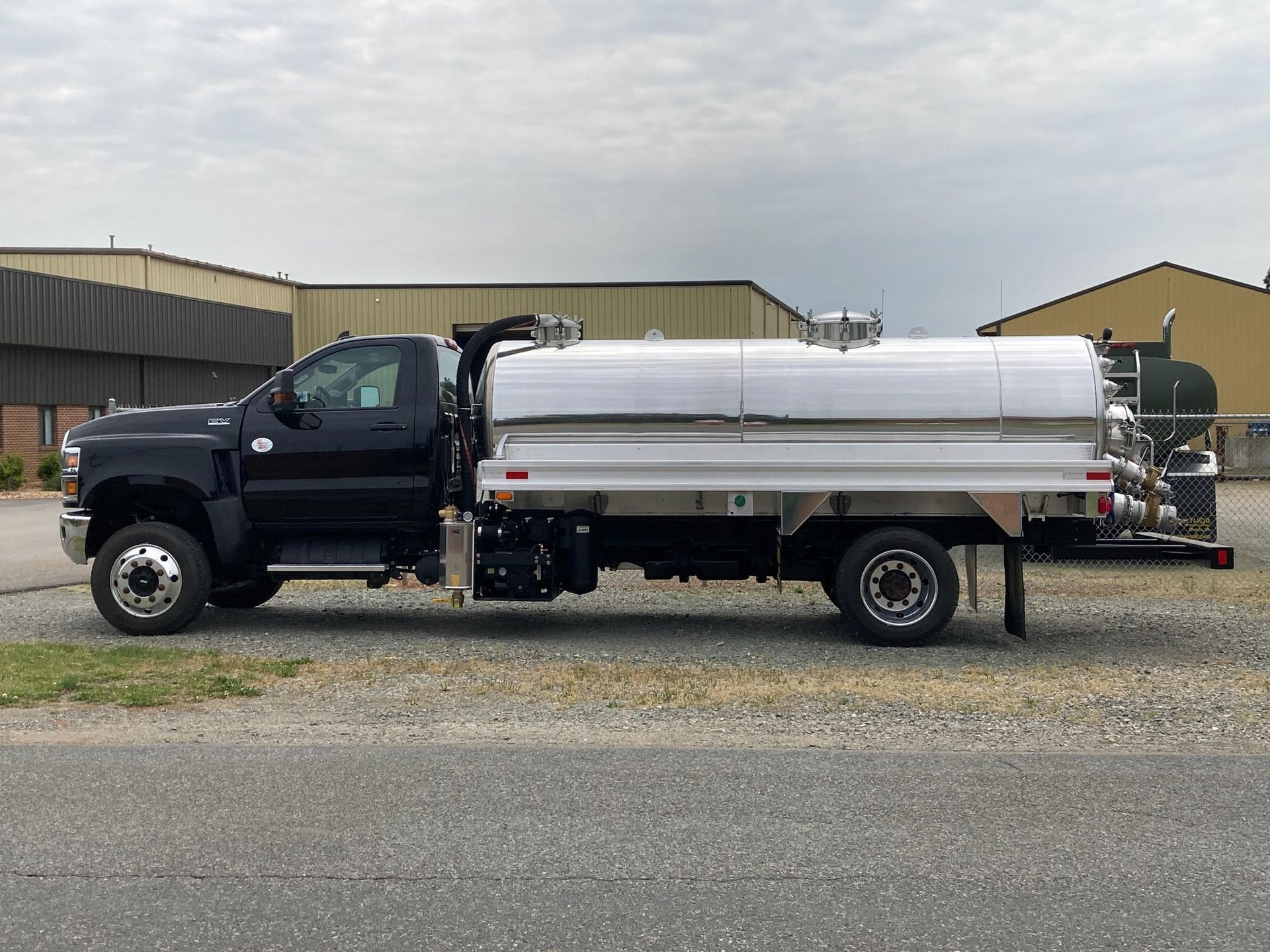 Black truck with large silver tank parked on gravel, cloudy sky.