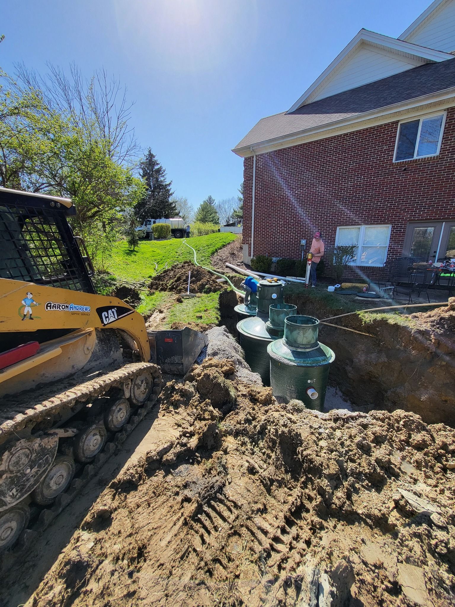 Skid steer and septic tanks being installed near a brick house on a sunny day.