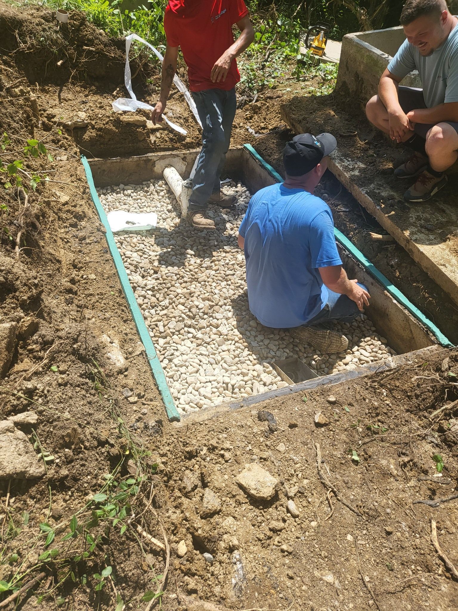 Three people installing a gravel-filled drainage system in an excavated area.