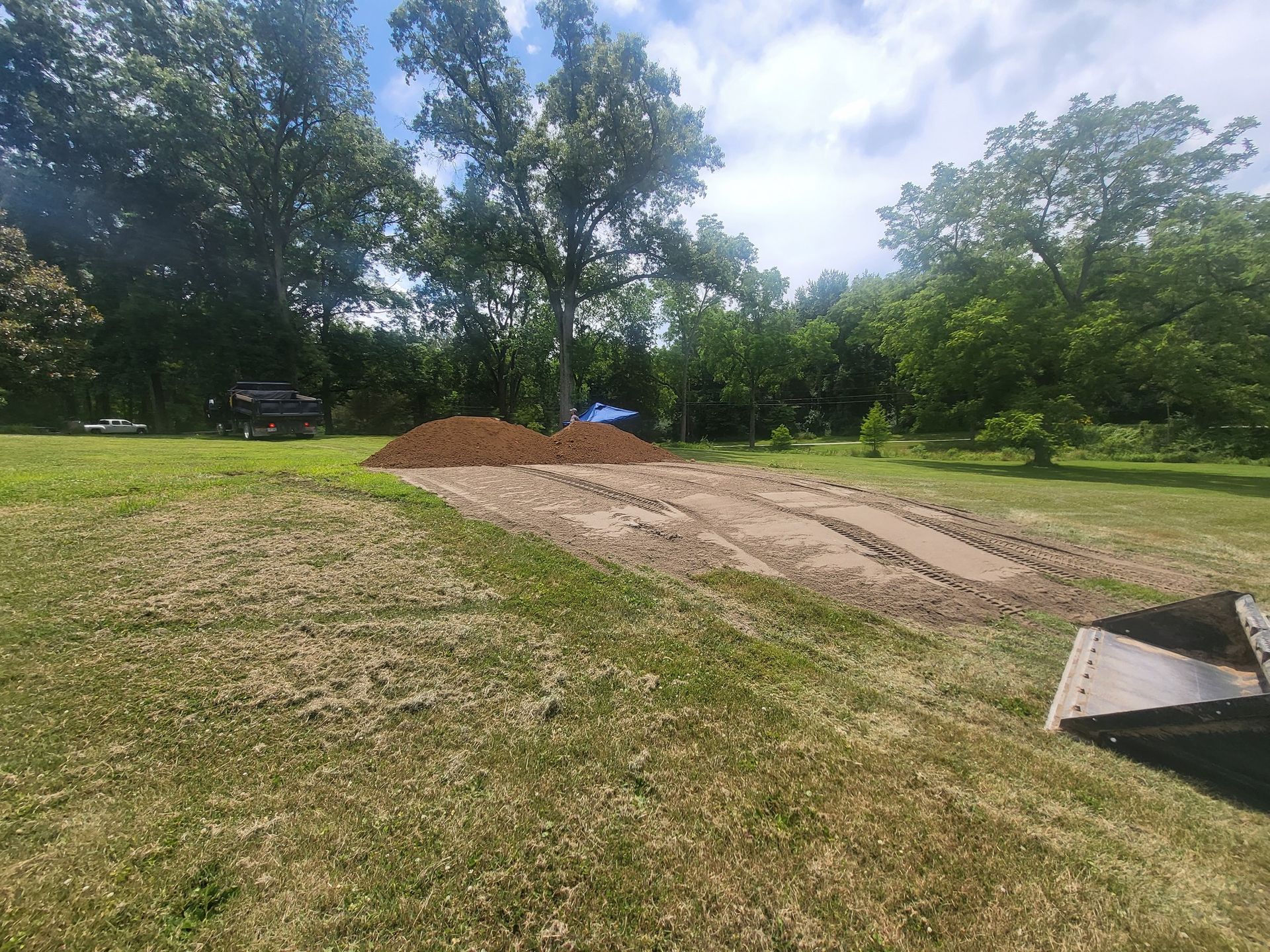 Dirt pile and prepared garden beds in a grassy yard, trees in the background, sunny day.