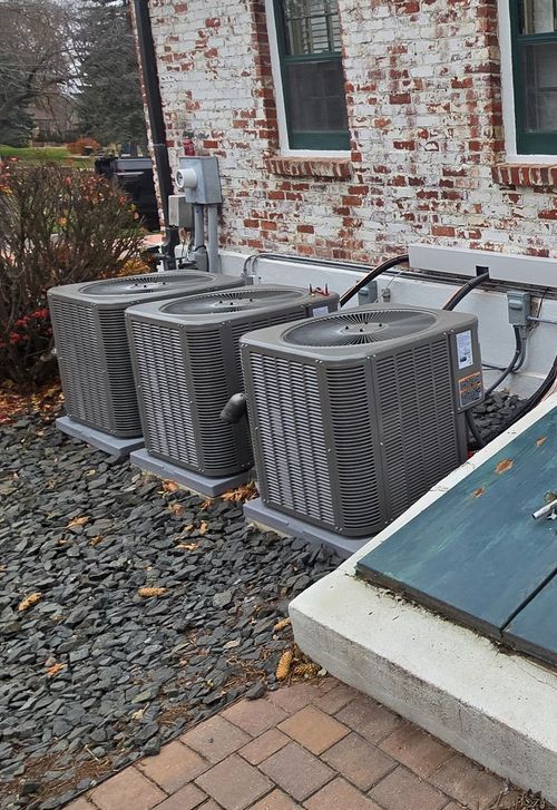 Two dark-colored air conditioning units on concrete pads next to a beige house, surrounded by gravel and grass.