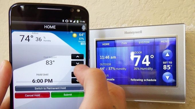 A person's hand using a smartphone app to adjust a Honeywell thermostat. The app shows 74° outside, and they're setting the thermostat to 83°.