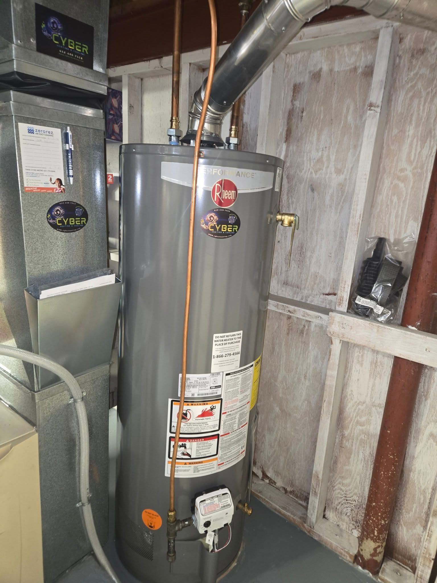 A tall, gray water heater in a basement next to a furnace and a wall.
