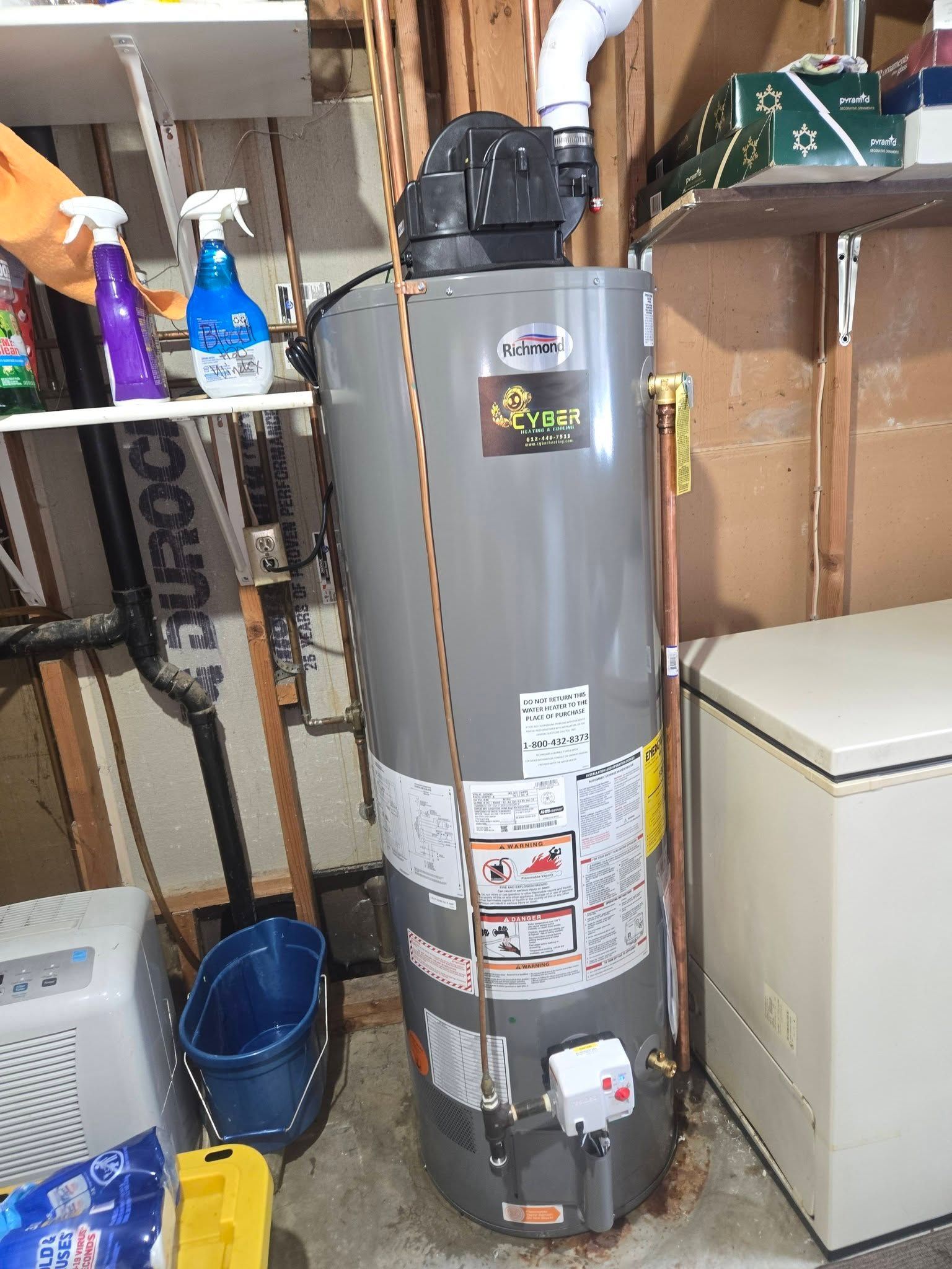 Gray water heater with copper pipes, vent, and control box in a basement near a freezer and shelf of cleaning supplies.