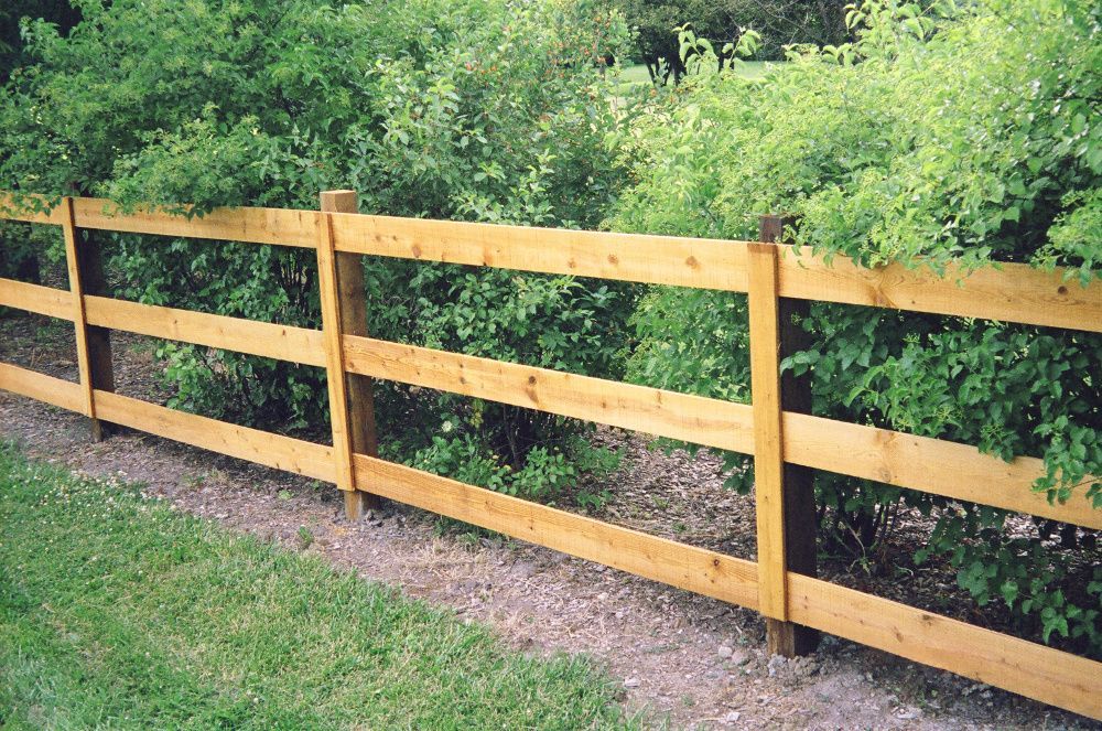 Wooden three-rail fence in front of green bushes and grass.