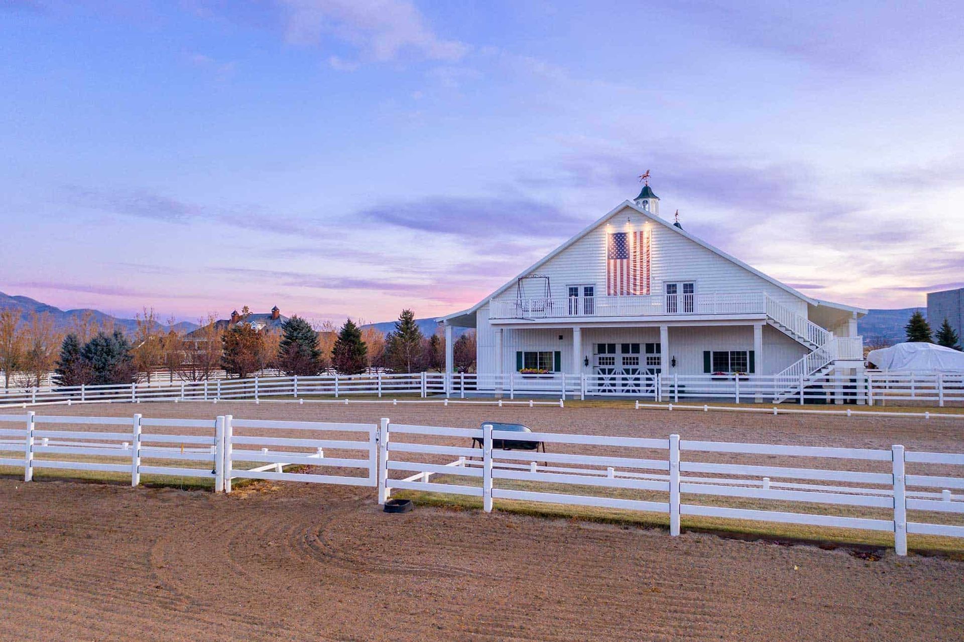 White barn with American flag at sunset, fenced equestrian area, mountains in background.