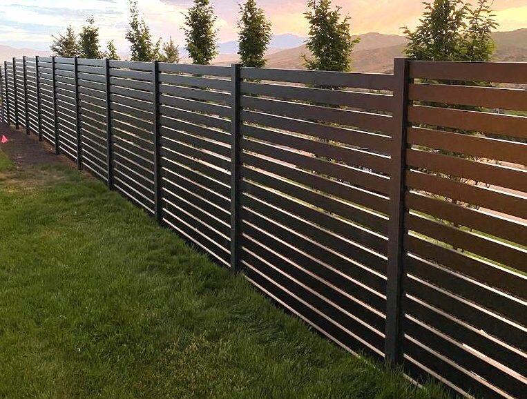 Dark horizontal slat fence along a green lawn with a tree line and mountains in the background.