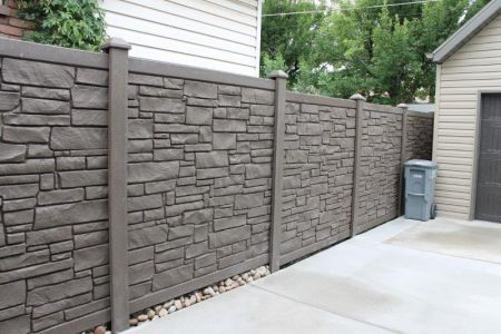 Stone-textured, brown privacy fence along a concrete driveway, near a building with a garage door and a green trash can.