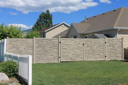 Stone textured fence in a backyard, with white picket fence on the left and house in the background. Blue sky overhead.