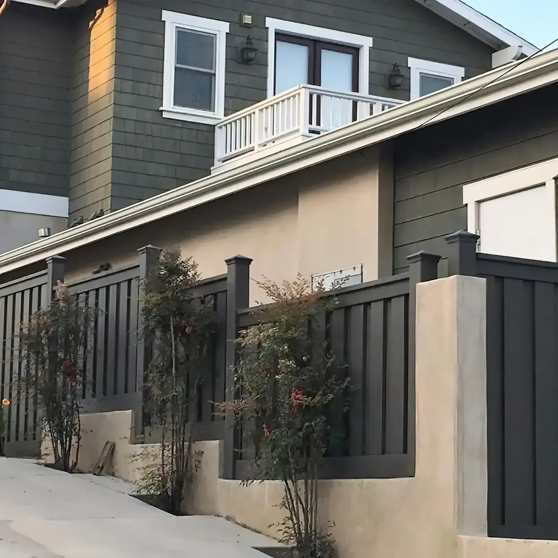 Dark gray fence and house with matching siding.  Tan stucco wall and sidewalk with small shrubs.