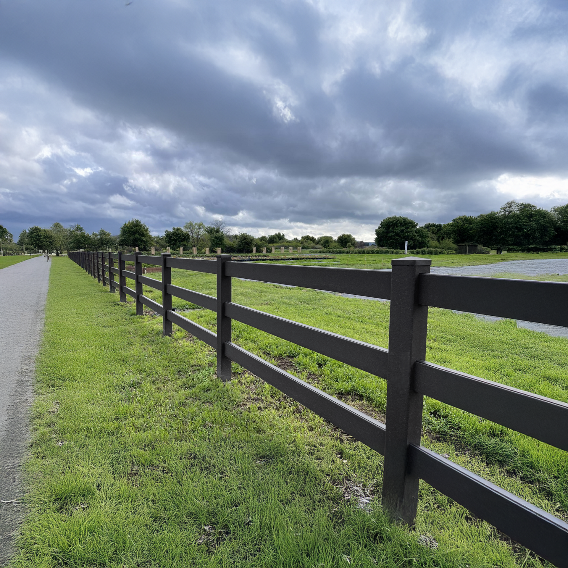 A dark brown fence borders a grassy area next to a paved path under a cloudy sky.