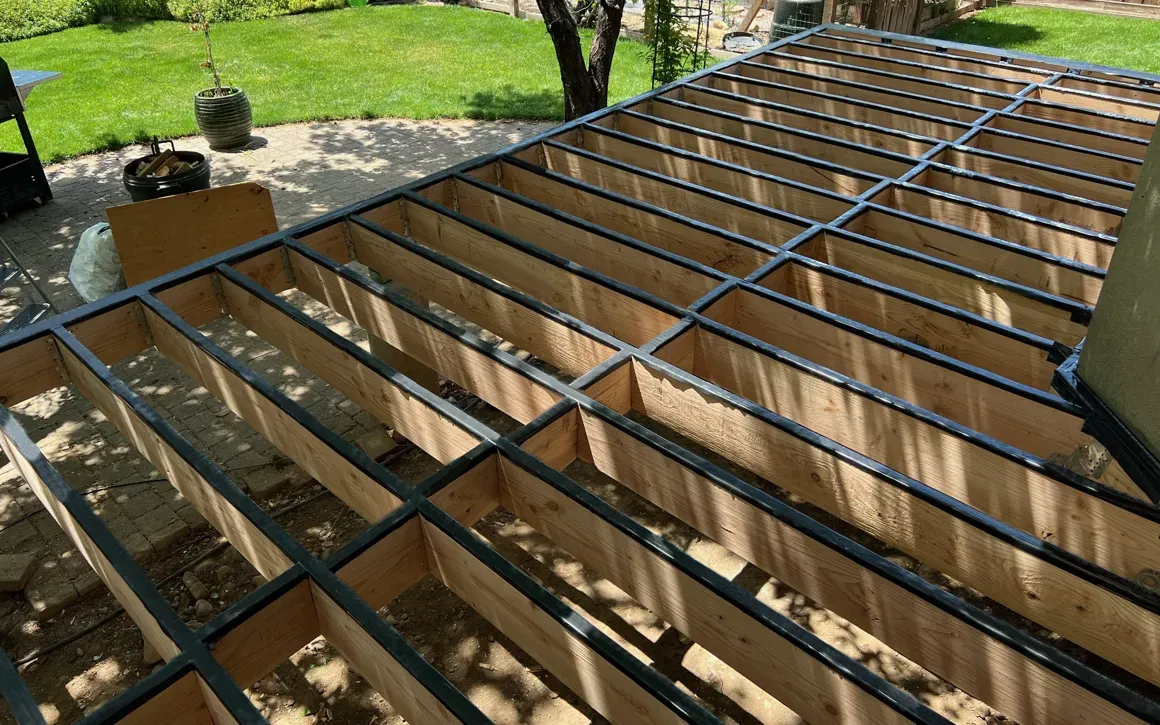 Wooden deck frame construction with black metal edging. Outdoors, sunny day.