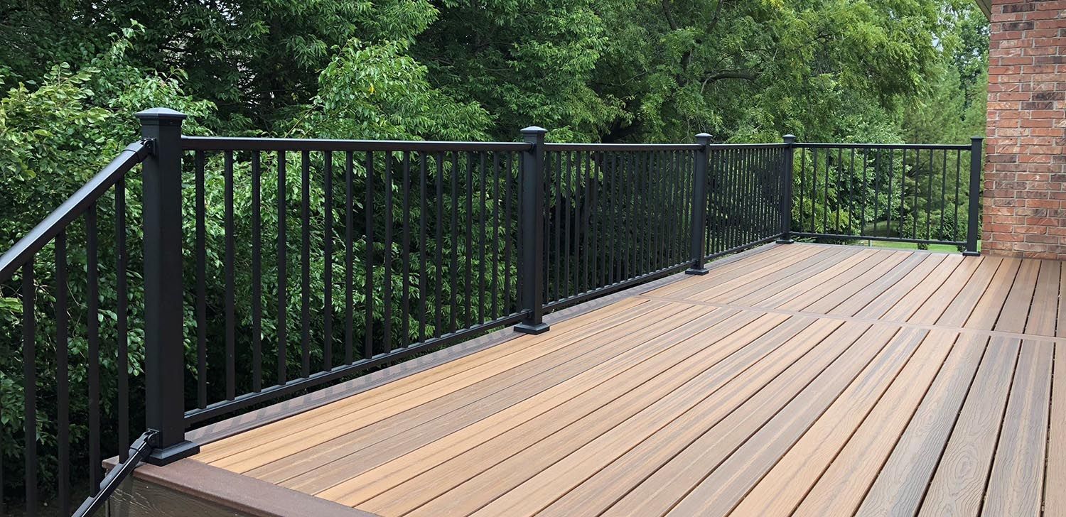 A wooden deck with black railings surrounded by lush green trees and a brick wall.