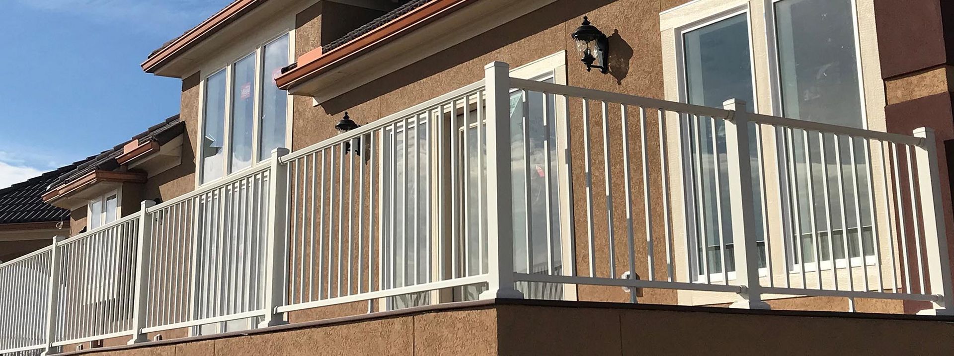 White railing on a balcony of a tan house with windows and a blue sky above.