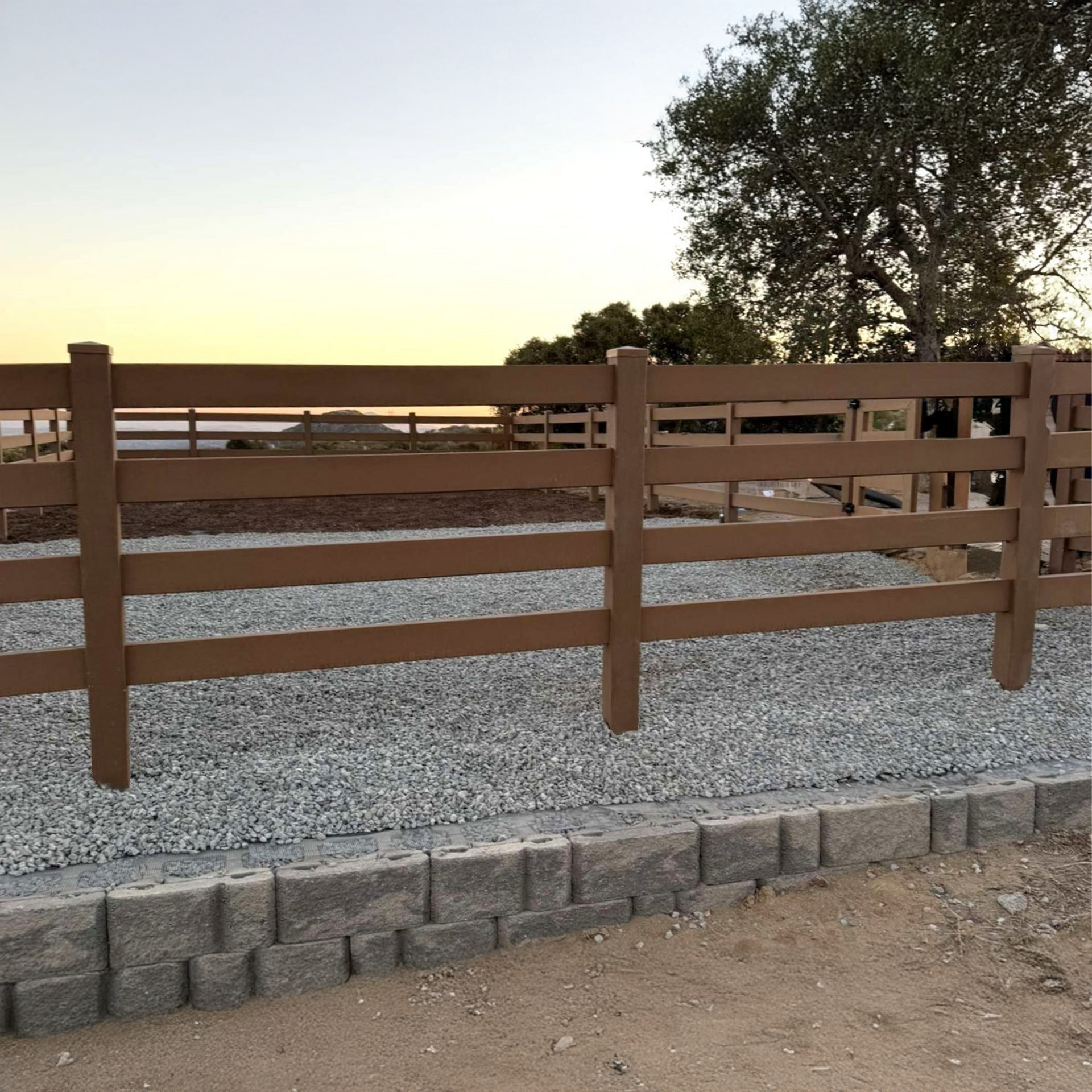 Brown wooden fence with gravel base against a pale sky and a tree.