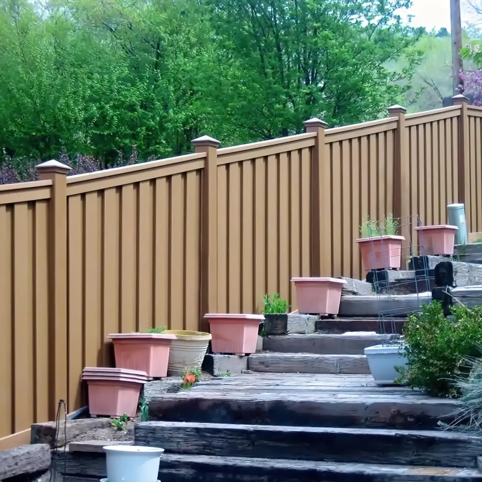 Brown fence, steps, and potted plants on a hillside, surrounded by greenery.