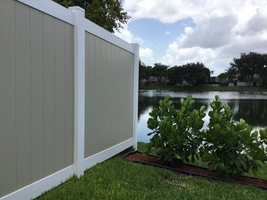 Tan and white privacy fence beside a lake, with green bushes and grass in the foreground.