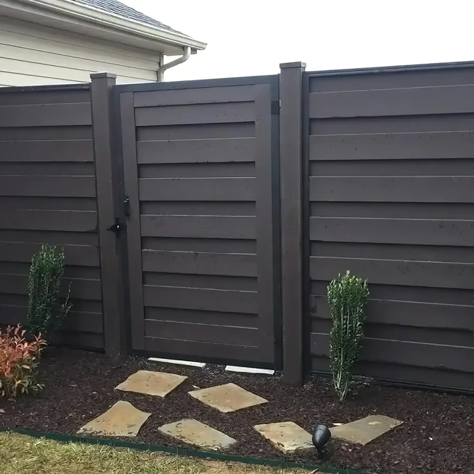 Brown horizontal slat fence with gate, stone path, and greenery.