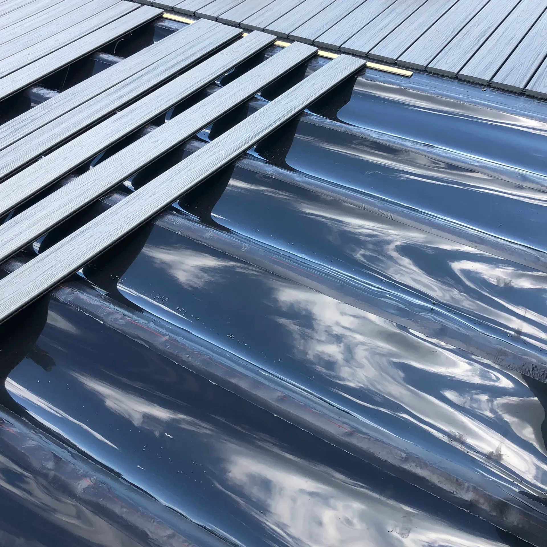 Gray deck boards partially covering a dark, corrugated sheet material, possibly roofing.