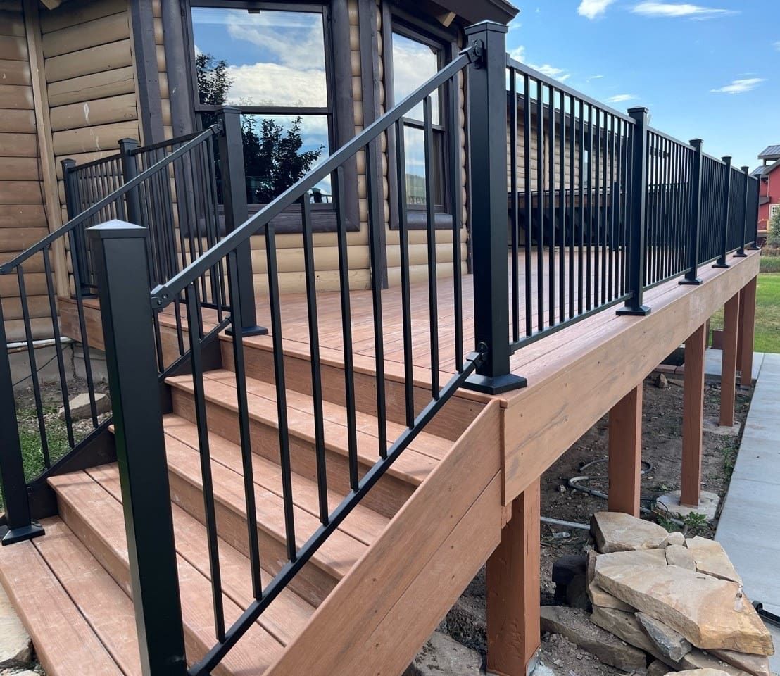 Black railing surrounds a wooden deck with stairs leading to a house with large windows.
