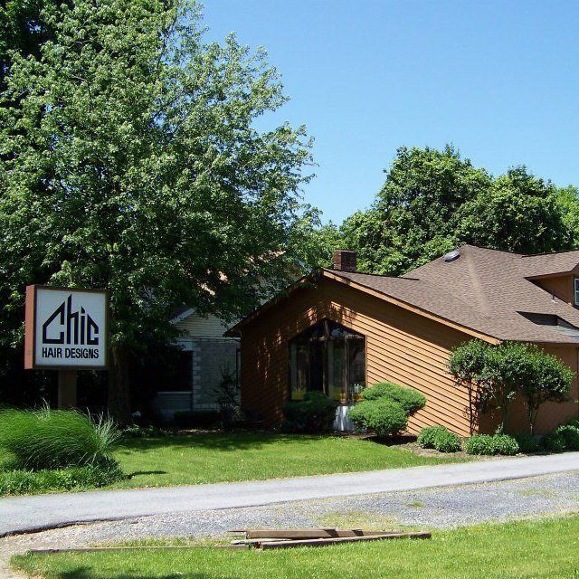 Sign for Chic Hair Designs in front of a brown building with a green lawn and trees.