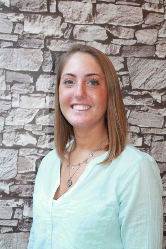 Woman with shoulder-length blonde hair smiles, wearing a light blue top, posing in front of a stone wall.