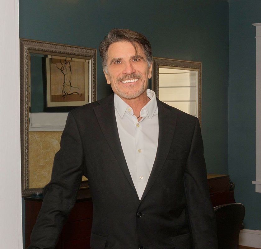 Man in black suit and open white shirt smiles, standing in a room with mirrors and art.
