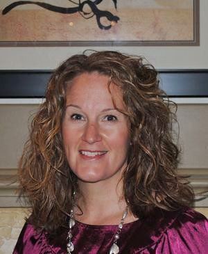 Woman with curly brown hair smiles, wearing a purple top and necklace, in front of art.