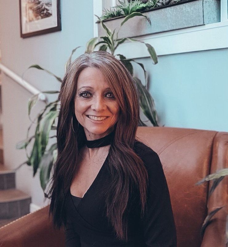 Woman with long brown hair smiling, sitting in brown chair, indoors.