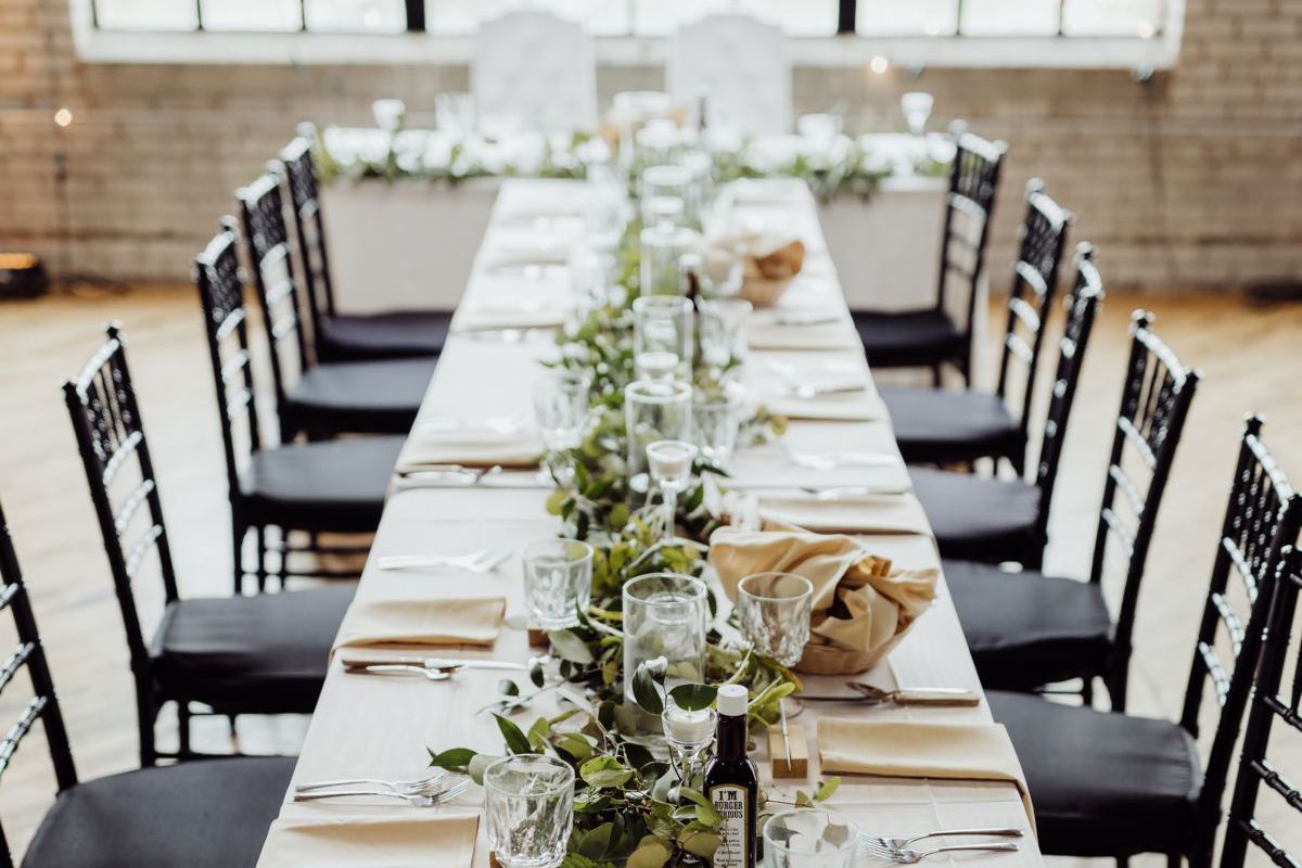 Long banquet table set with white linens, black chairs, and floral centerpieces in a bright dining room