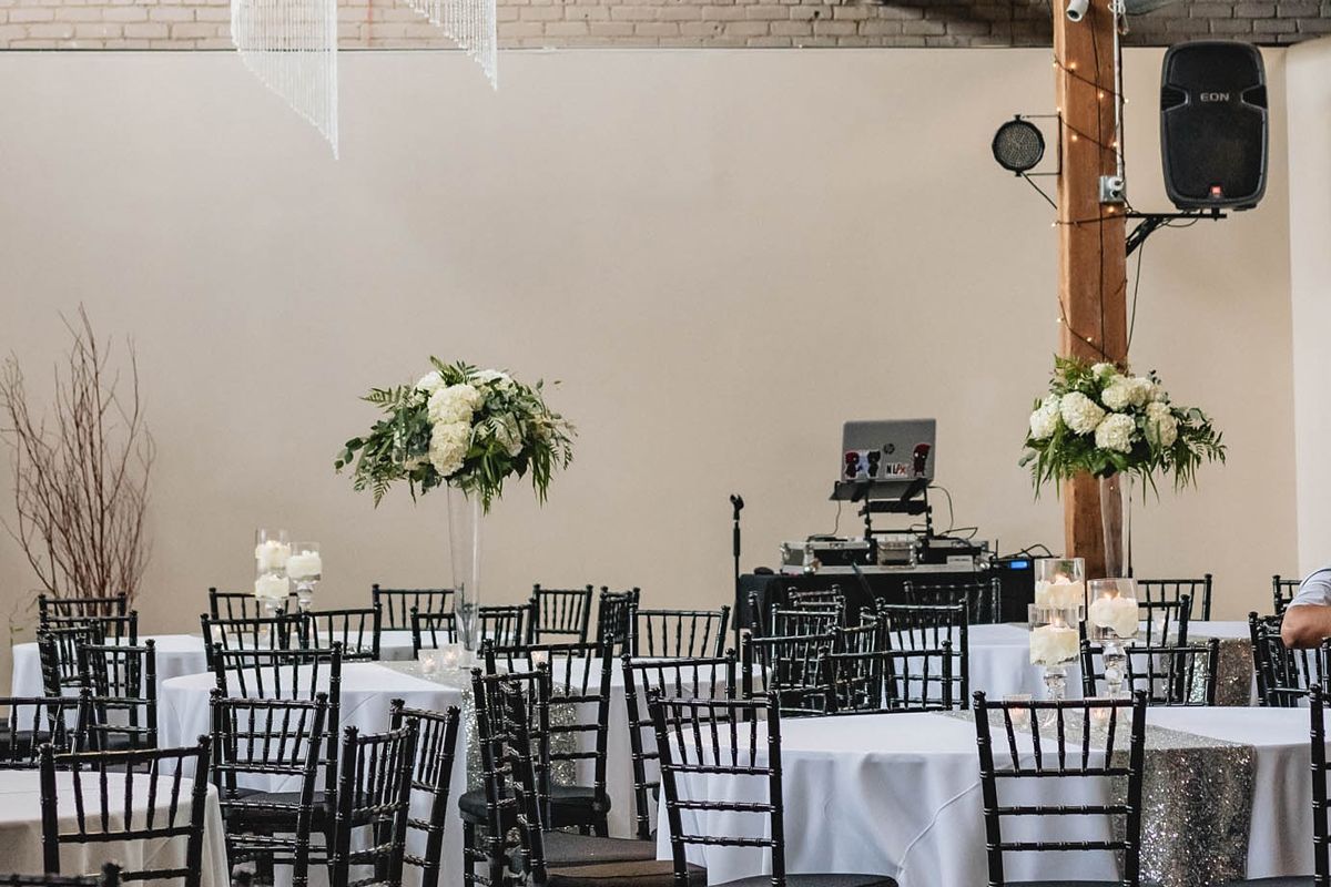 Elegant event hall with round tables, white linens, black chairs, and floral centerpieces under a wood beam.