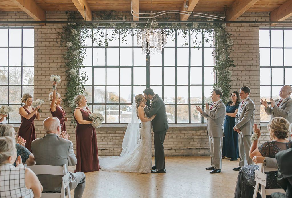 Wedding couple kissing under floral arch in bright industrial venue with guests applauding