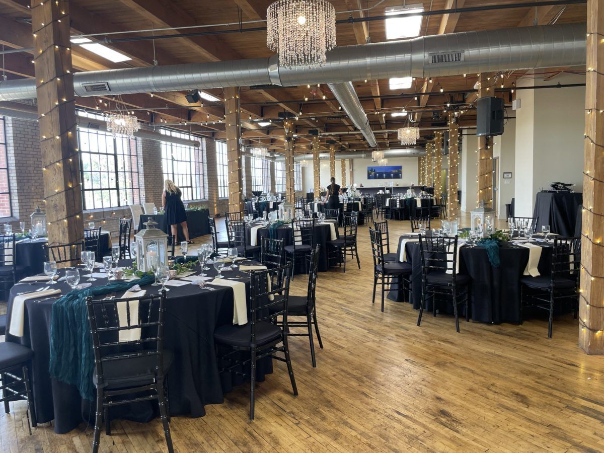 Empty banquet hall with black tablecloths, chairs, and chandelier lighting in a wooden-floored loft space