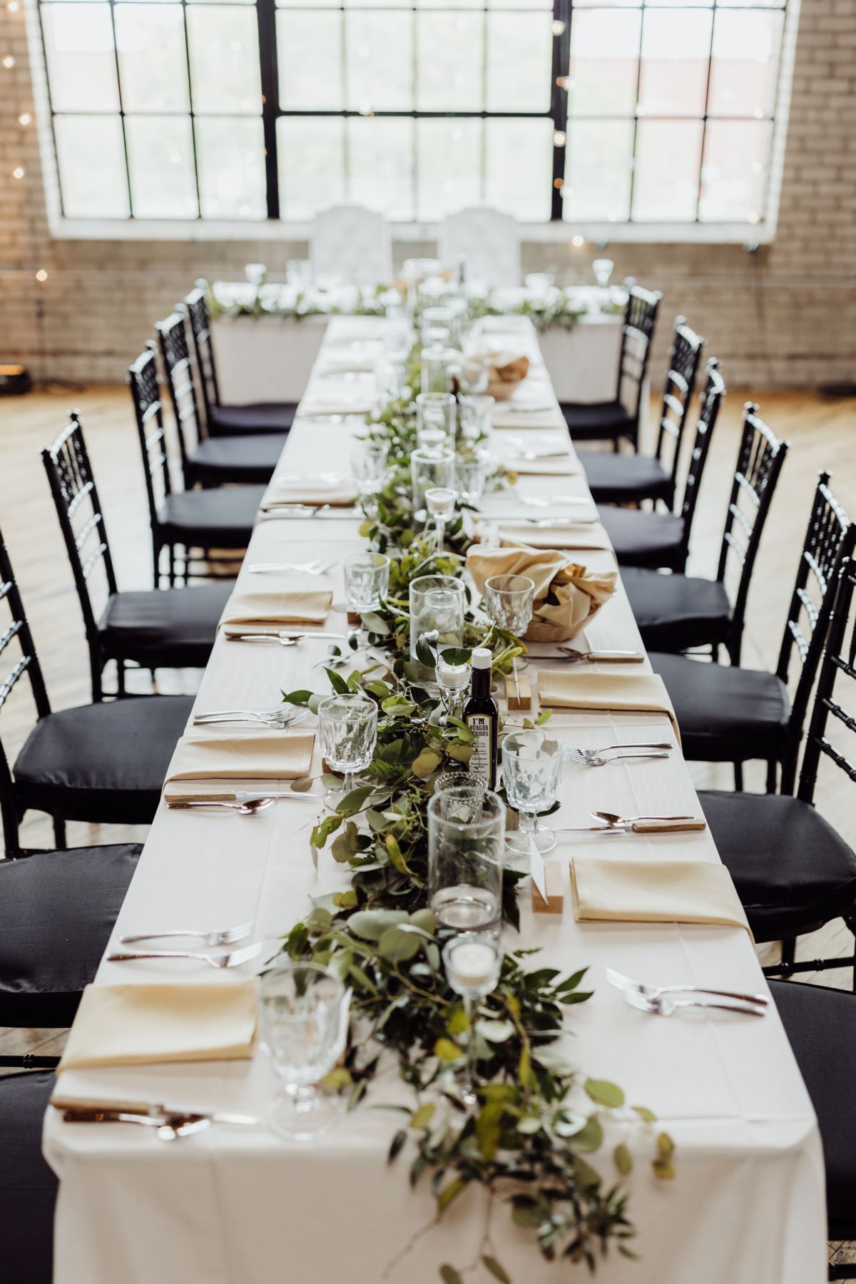 Long banquet table with white linens, black chairs, and greenery centerpiece in a bright industrial room