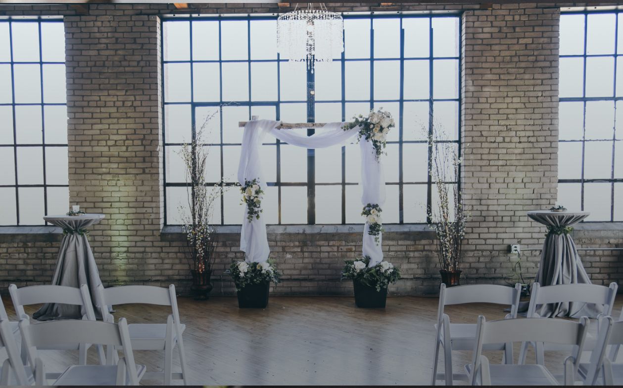 Wedding ceremony setup: Wooden arch with white fabric and flowers, against a large window, white chairs, and gray tablecloth tables.