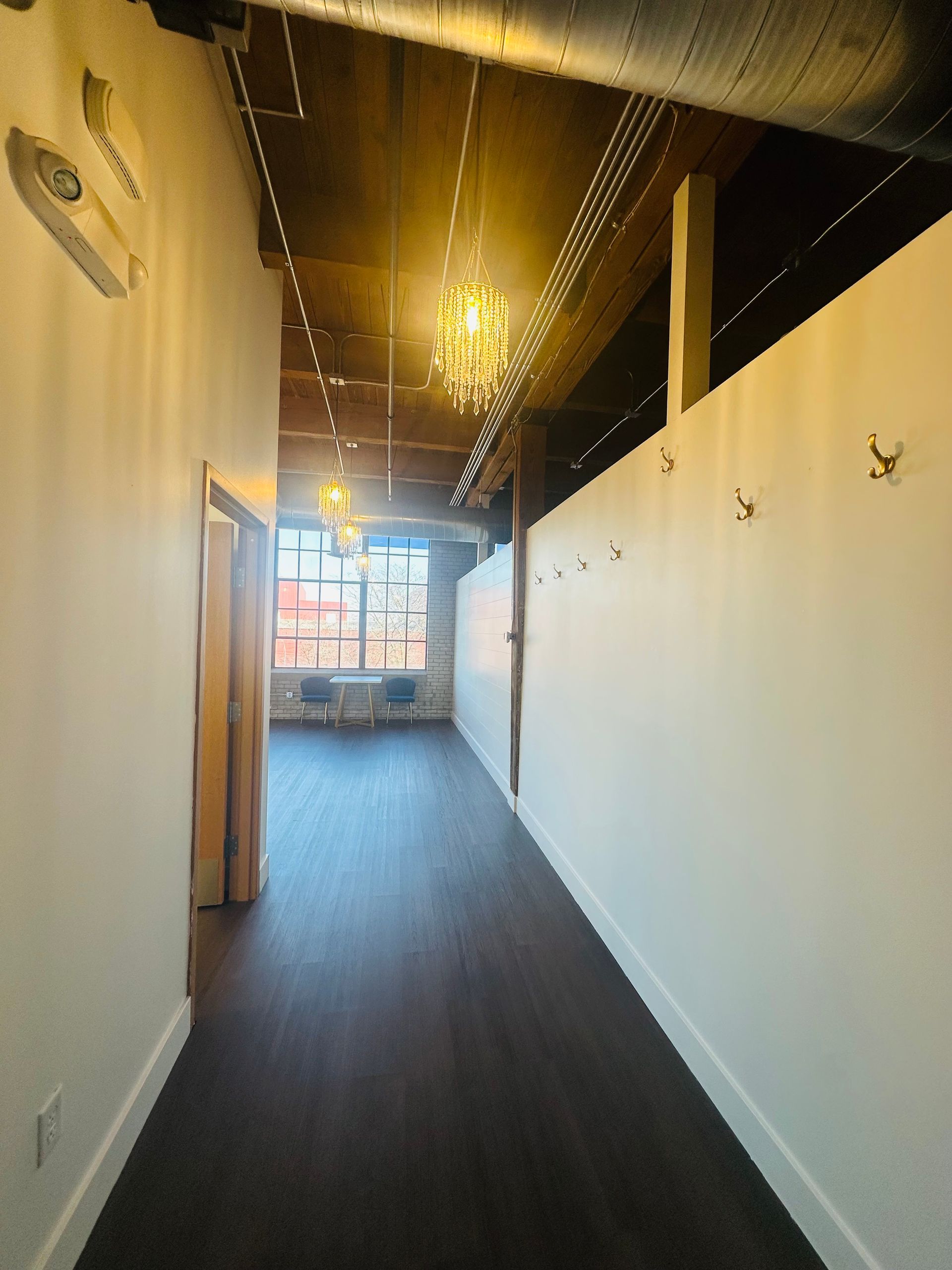 A hallway with dark wood floors, white walls with wall-mounted hooks, exposed ceiling beams, and a bright window at the end.