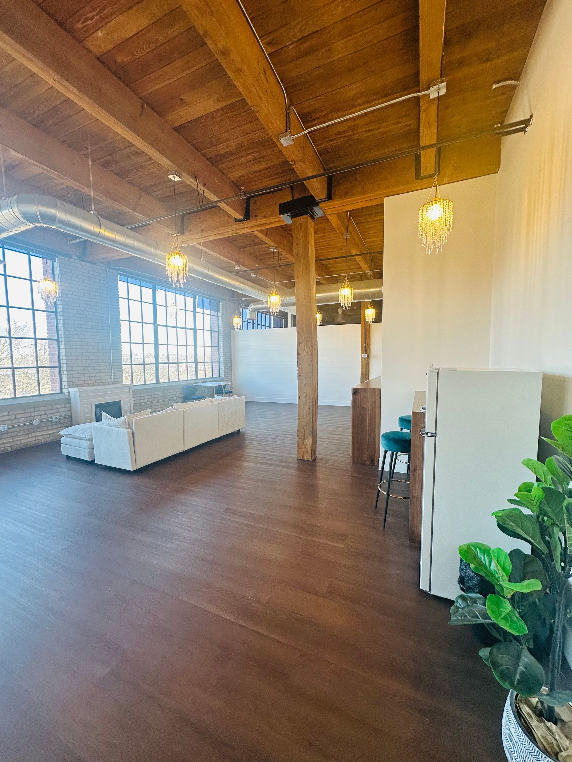 An open-concept loft with a white sectional, wood-beamed ceiling, tall windows, and a plant in the foreground.