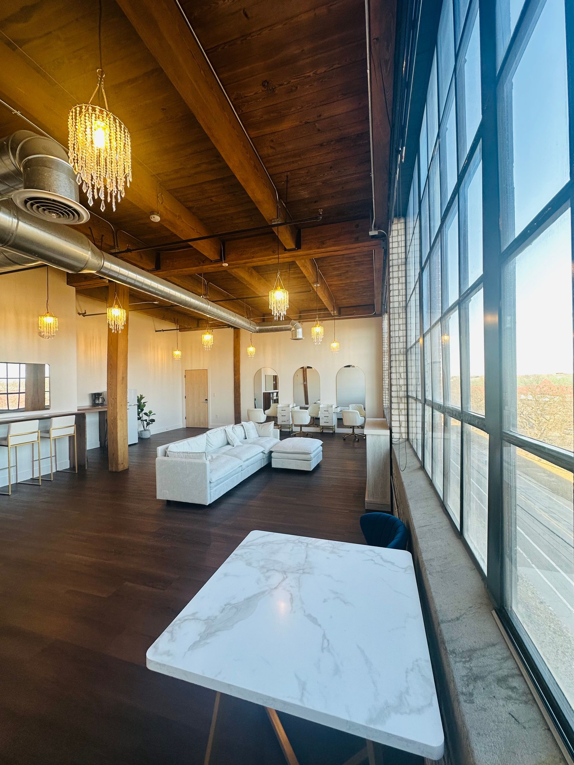 A spacious industrial-style loft featuring exposed wood beam ceilings, large windows, a white sofa, and a kitchen area.