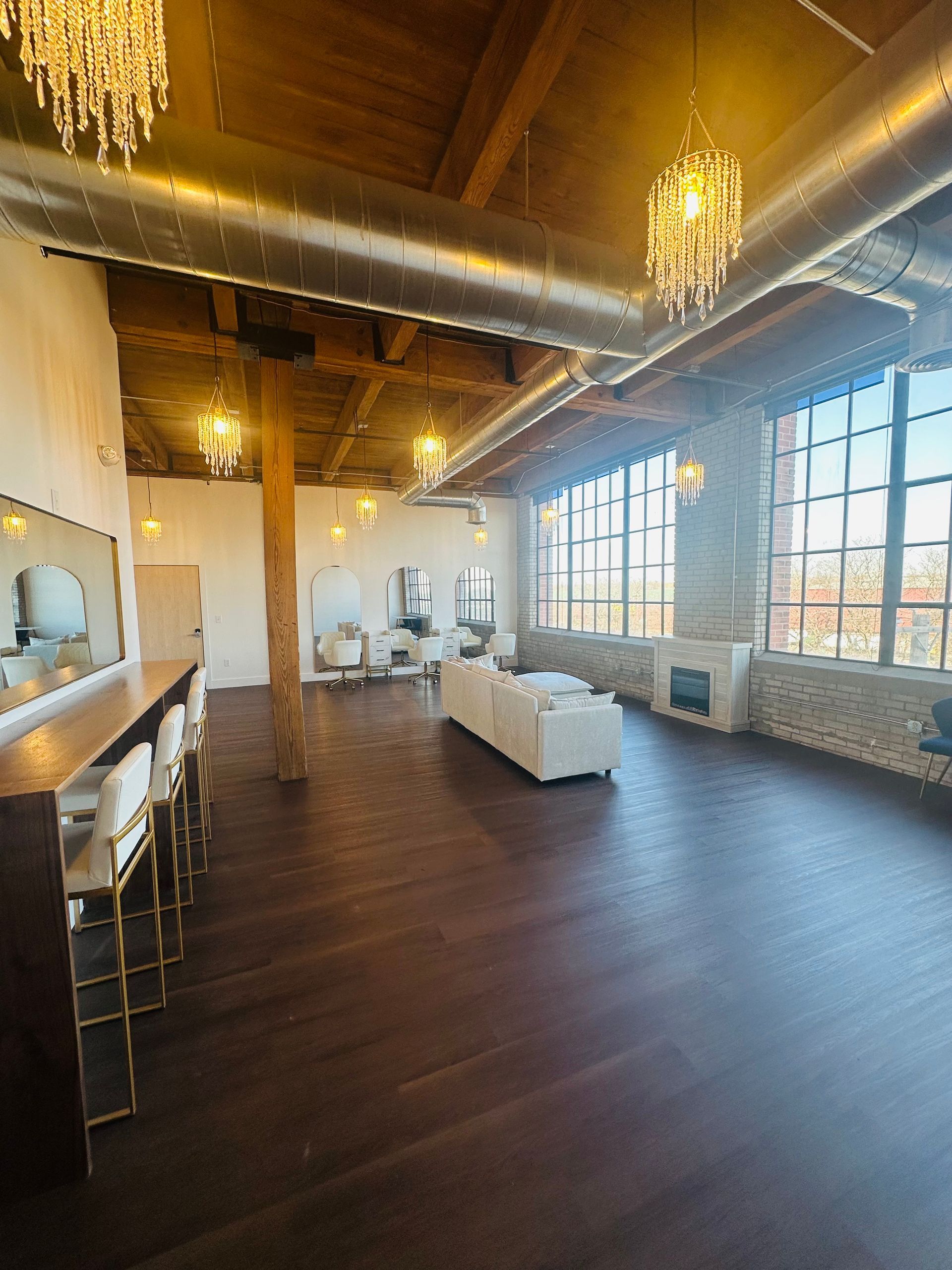 An open loft salon featuring dark wood floors, white seating, large windows, and crystal chandeliers under wooden beams.