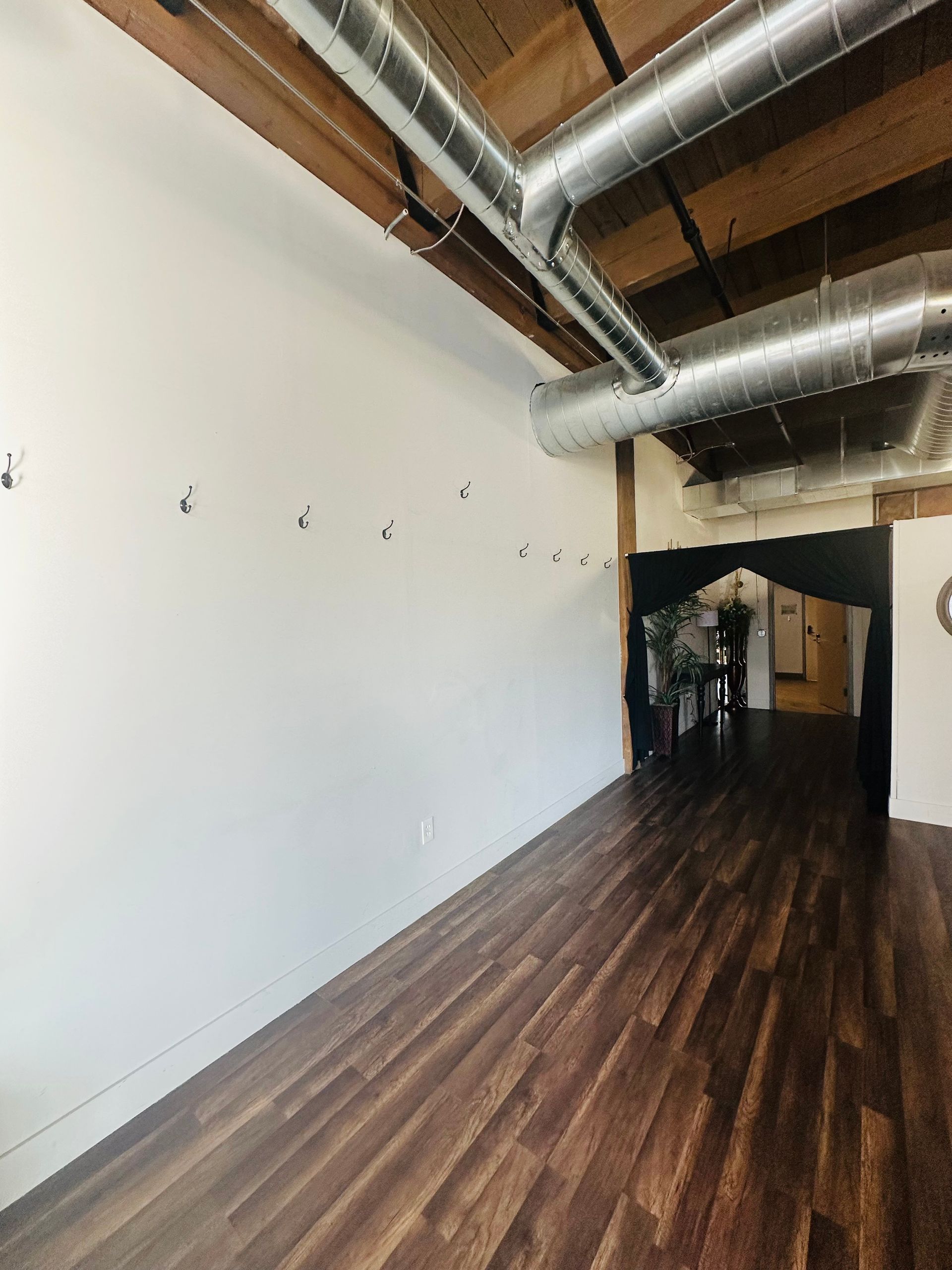 Empty room with dark wood flooring, white walls with small hooks, and exposed industrial ceiling pipes.