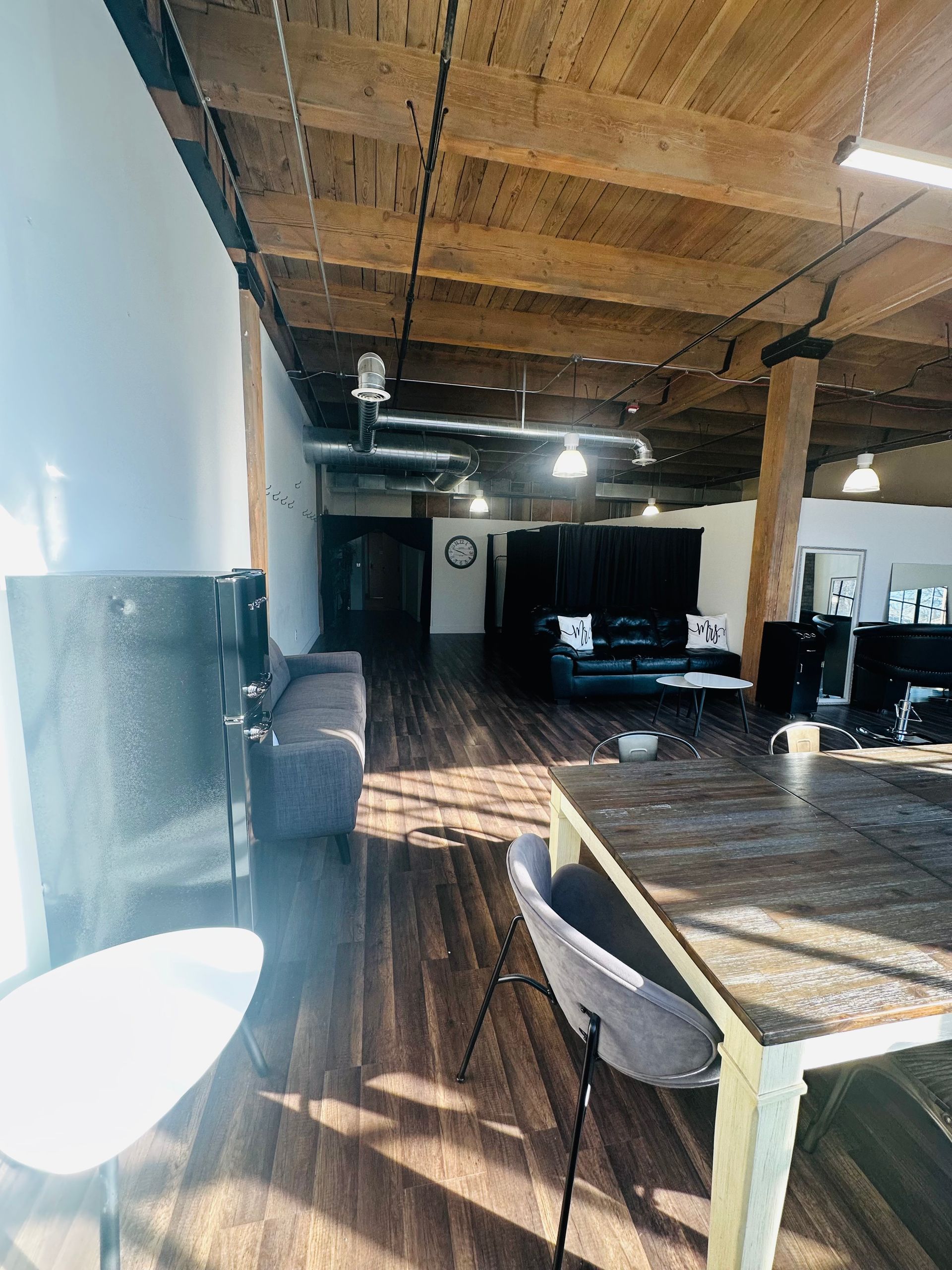 An open-plan loft office with exposed wooden beams, dark wood floors, a table, gray chairs, and a couch in the distance.
